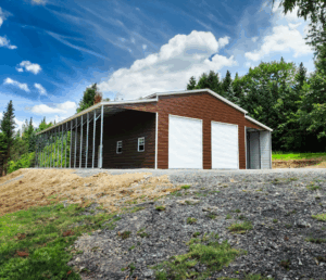 Spacious brown metal barn with white trim and dual roll-up garage doors, featuring an open-sided extension for added storage—perfect for use as a barn, farm equipment, or workshop space in a rural setting.